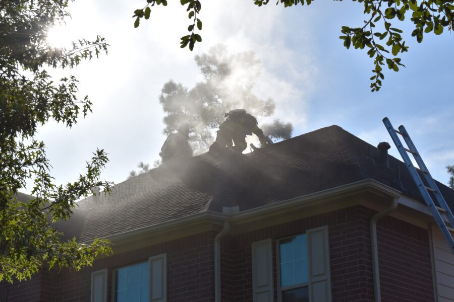 Two firefighters stand on a smoky residential roof, working to ventilate the attic as sunlight shines through surrounding trees.