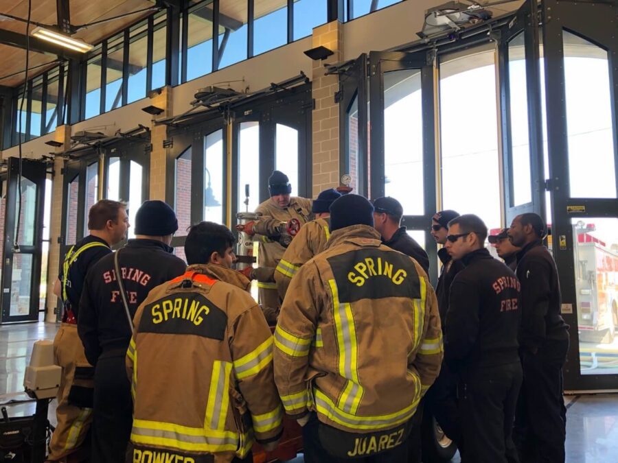 A group of firefighters in uniform, labeled "Spring Fire," stand in a fire station near open garage doors, gathered around equipment.