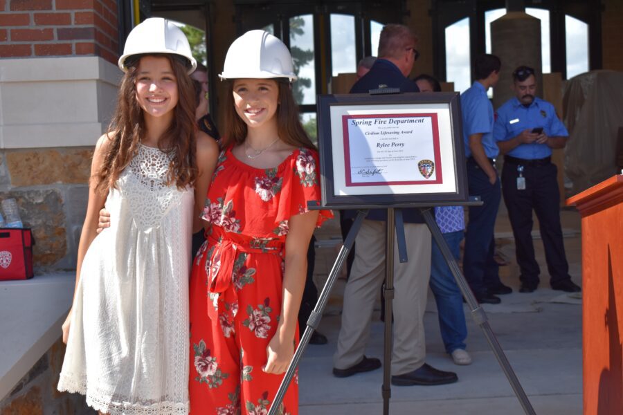 Two young women wearing white hard hats stand smiling next to an award display at a public event, with other people and a fire department certificate in the background.