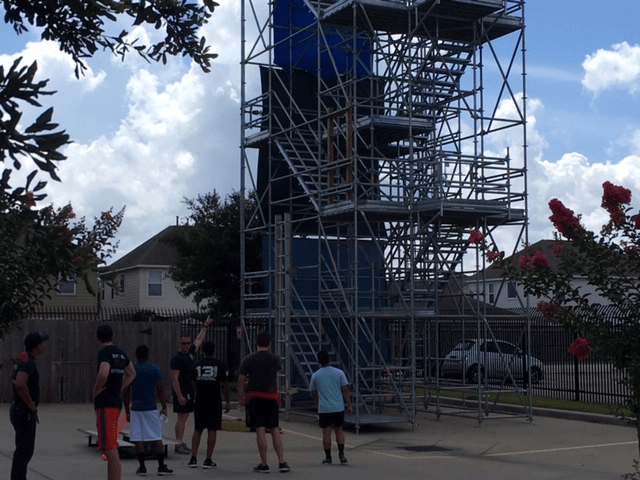 A group of people stand in front of a tall metal scaffolding structure outdoors, with houses and trees in the background.