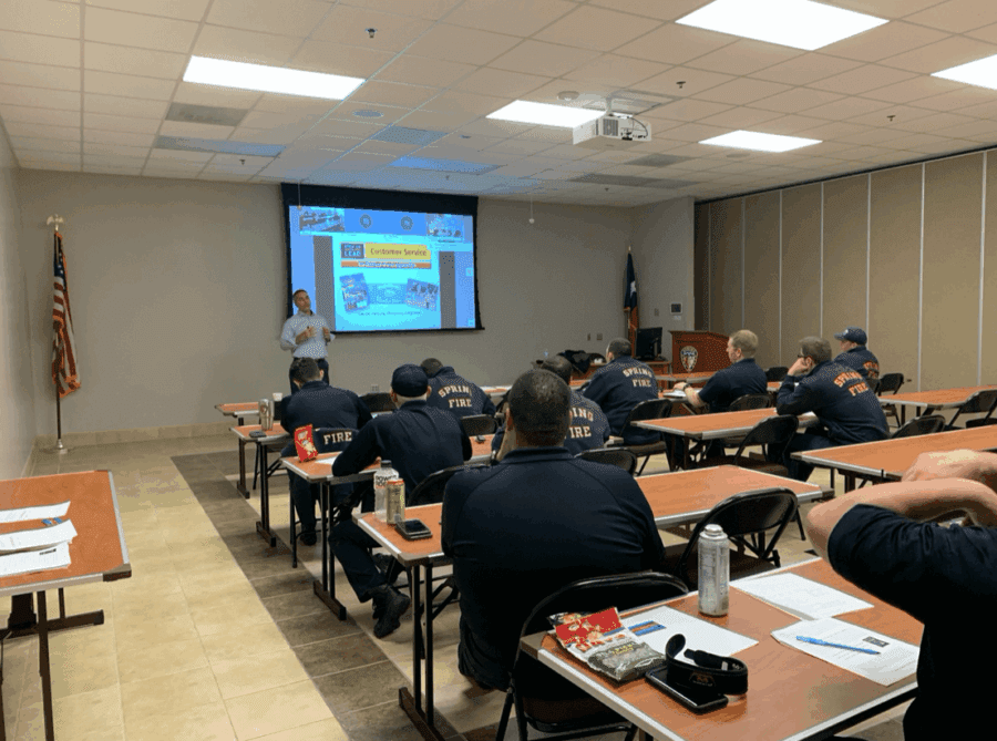 A group of firefighters in uniform sit at tables in a classroom, listening to a presentation with slides projected on a screen at the front of the room.
