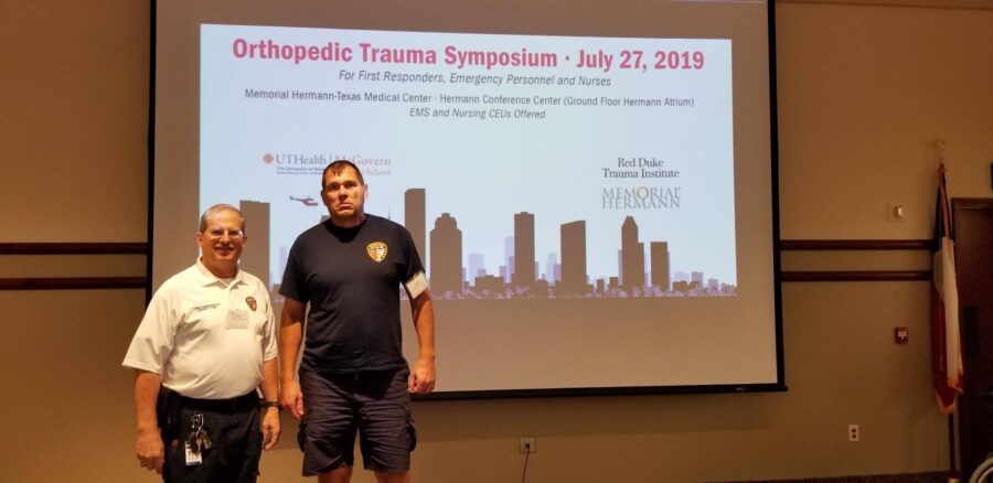Two men stand in front of a projector screen displaying details for the Orthopedic Trauma Symposium held on July 27, 2019, at Memorial Hermann-Texas Medical Center.