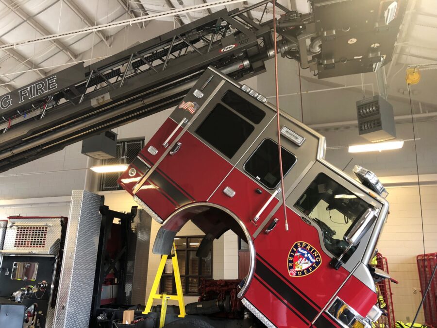 A red fire truck cab is tilted forward and propped up on a yellow stand inside a fire station, exposing the engine for maintenance.