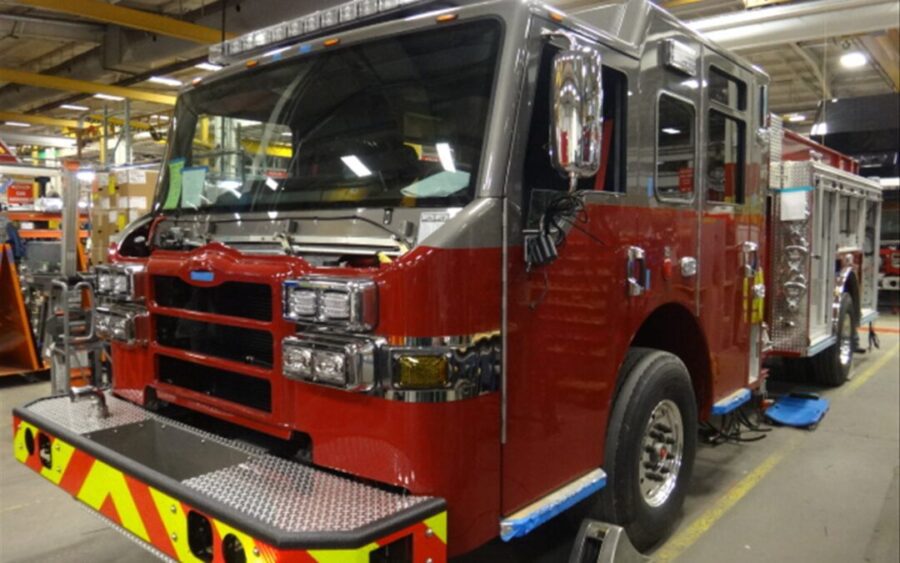 A red fire truck parked inside a large industrial garage, surrounded by tools and equipment, with work in progress on its exterior.