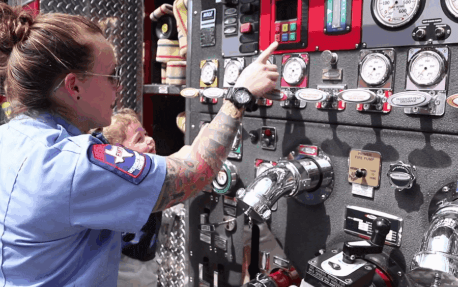 A firefighter in uniform points to control panels and gauges on a fire truck, with a child looking on in the background.