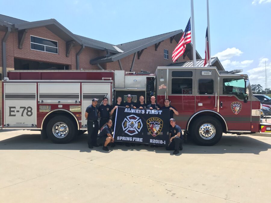 A group of firefighters poses in front of a fire truck holding a banner that reads "Always First Spring Fire" outside a fire station with flags in the background.
