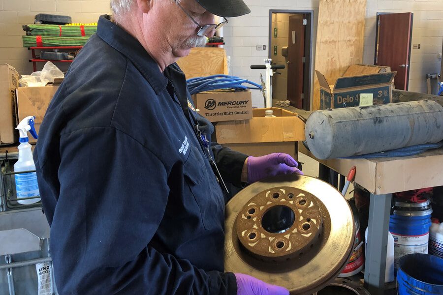 A person wearing gloves examines a circular metal part in a workshop with various tools and equipment in the background.