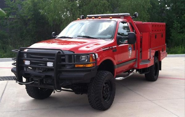 A red fire department utility truck with black grille guard is parked on a concrete surface, surrounded by trees.