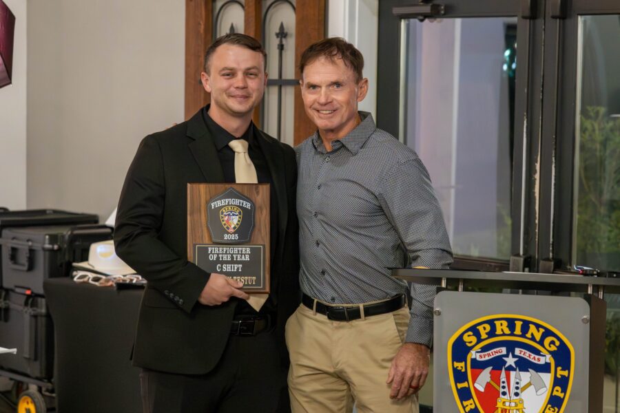 Two men pose indoors; one holds a "Firefighter of the Year" plaque. A podium with the Spring Fire Dept. logo is visible in the foreground.