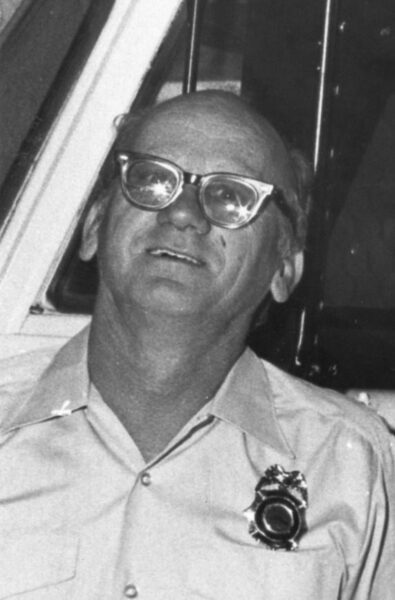 A man wearing glasses and a button-up shirt with a fire badge, standing in front of a vehicle.