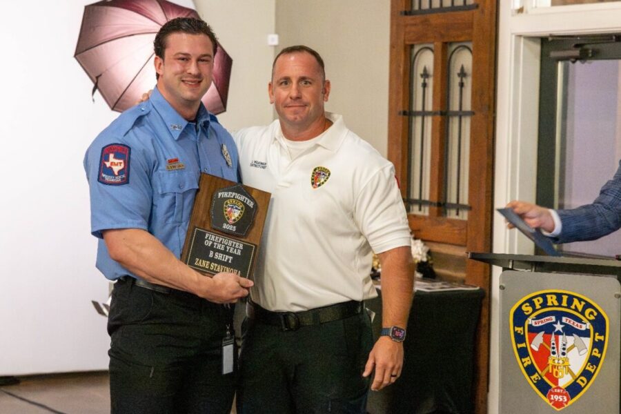 Two firefighters stand together indoors, one holding a "Firefighter of the Year" plaque. A Spring Fire Department emblem is visible on a podium beside them.