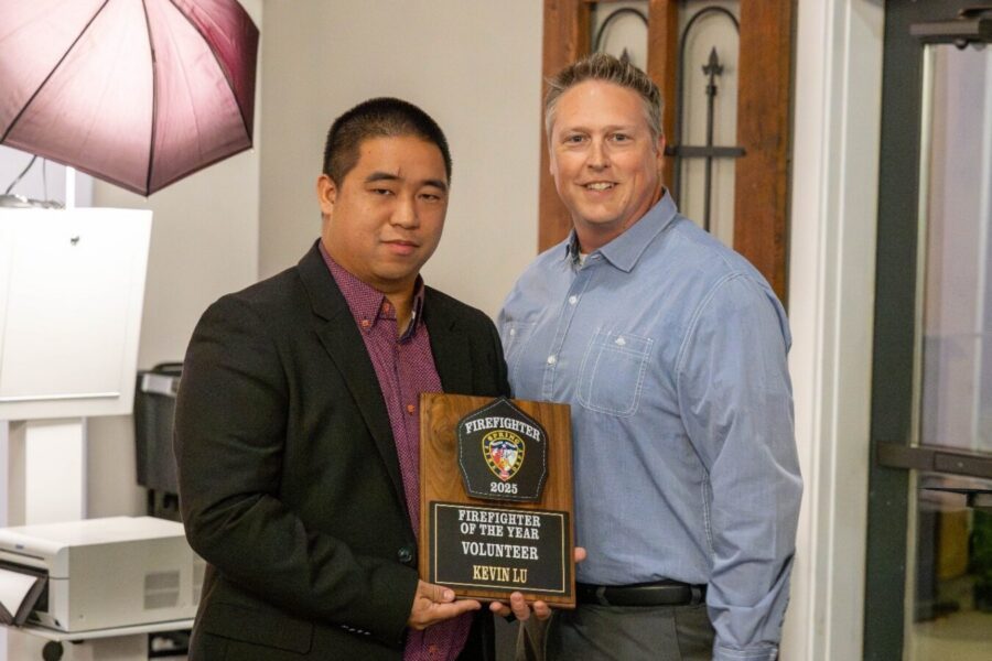 Two men stand indoors; one holds a plaque reading "Firefighter of the Year Volunteer 2023 Kevin Liu." Both are looking at the camera and smiling.
