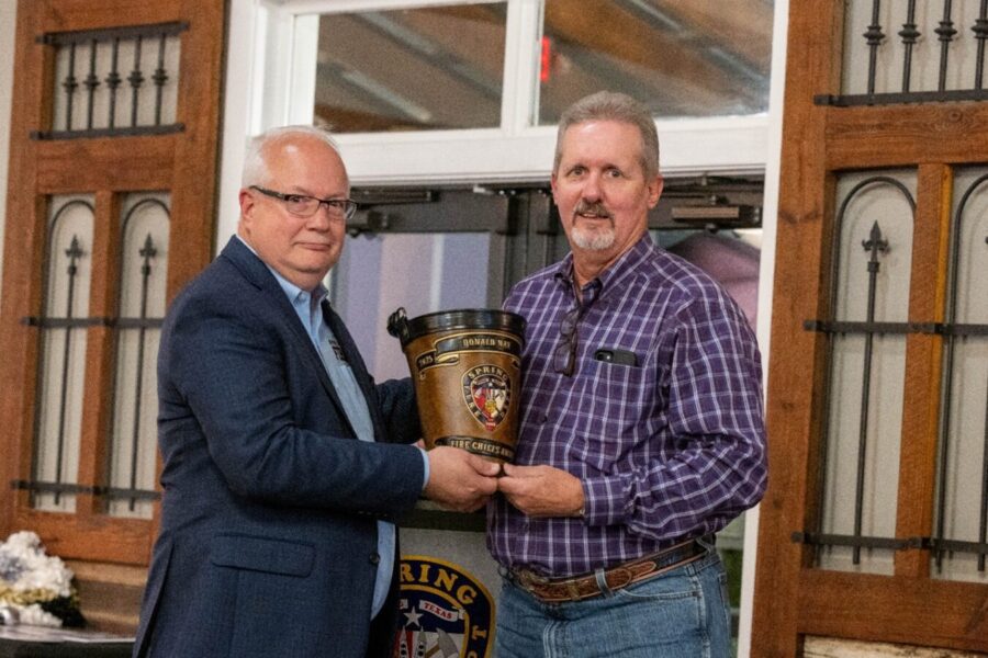 Two men stand indoors, one in a suit and the other in a plaid shirt, holding a commemorative fire department bucket together and posing for a photo.
