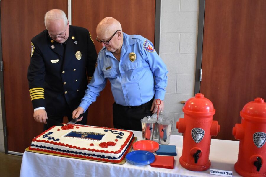 Two uniformed firefighters cut a decorated sheet cake on a table with red fire hydrant-shaped drink dispensers at each end.