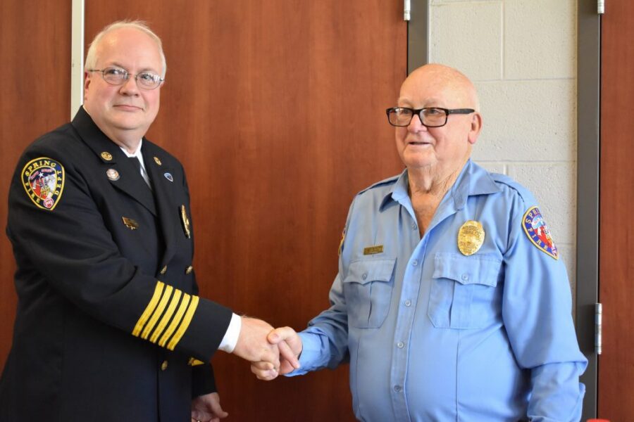 Two uniformed men, one in a black dress uniform and the other in a blue shirt, shake hands in front of a wooden door and gray wall.