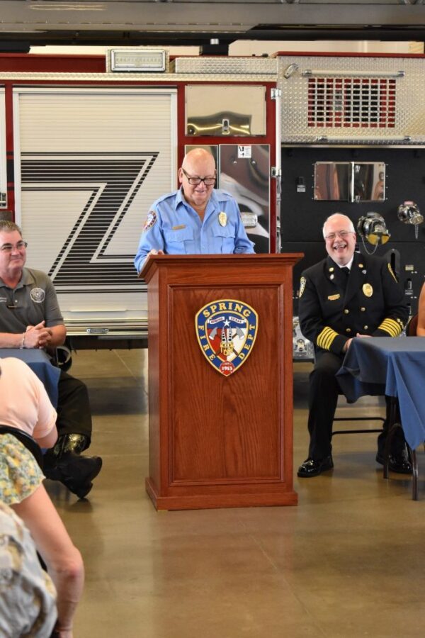 A man in a blue uniform speaks at a podium with a fire department emblem, flanked by officials in uniform, in front of a fire truck.