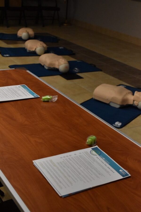 CPR training mannequins are set up on mats in a row behind a table with instruction sheets and earplugs in a classroom setting.