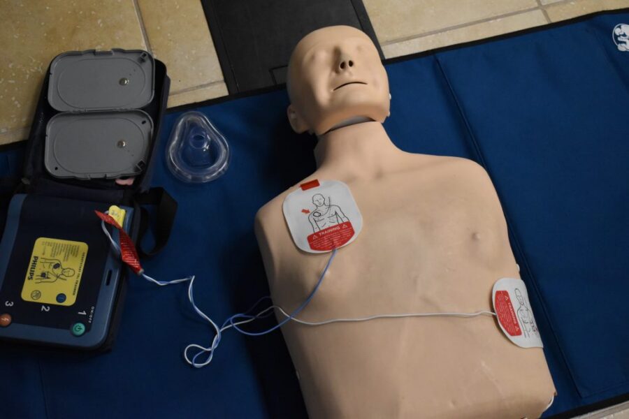 A CPR training manikin with AED pads attached to its chest, next to an automated external defibrillator and a face mask on a blue mat.